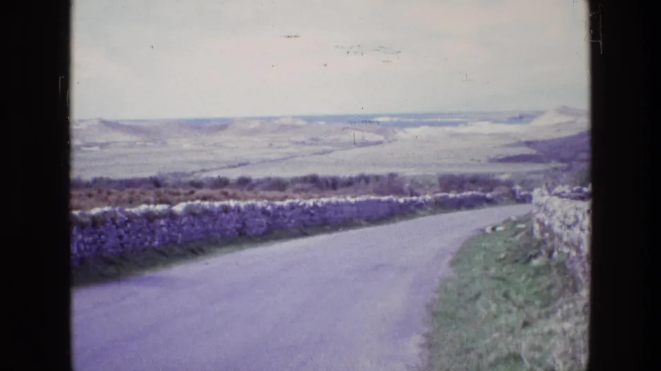 Final 1969 Ireland frame showing the same rural road curving past dry-stone walls toward distant rolling hills.