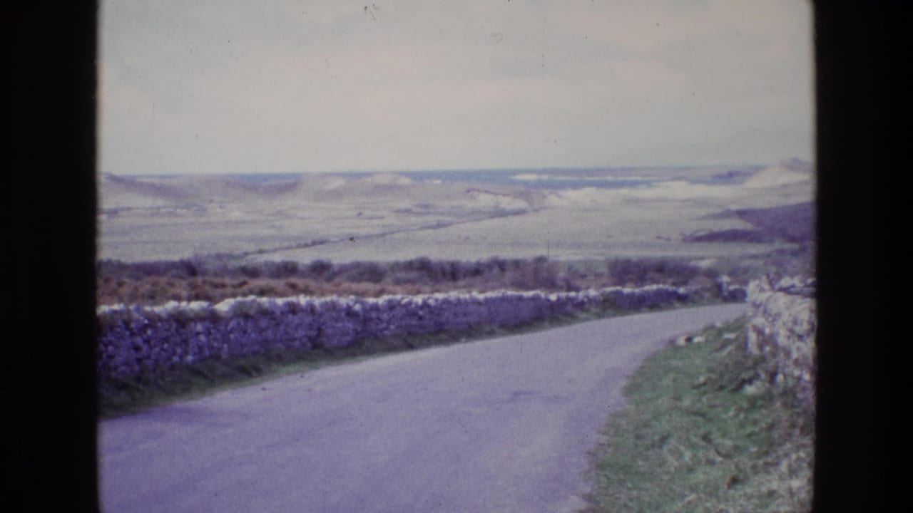 1969 Ireland home-movie view of a narrow road curving past stone walls toward open rolling hills under an overcast sky.