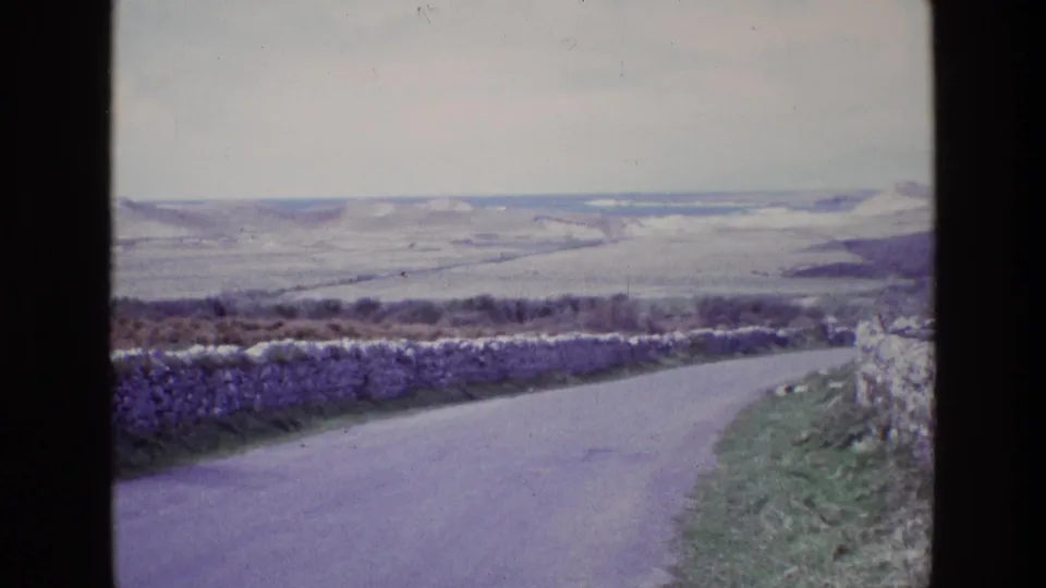 1969 Ireland home-movie view of a narrow road curving past stone walls toward open rolling hills under an overcast sky.