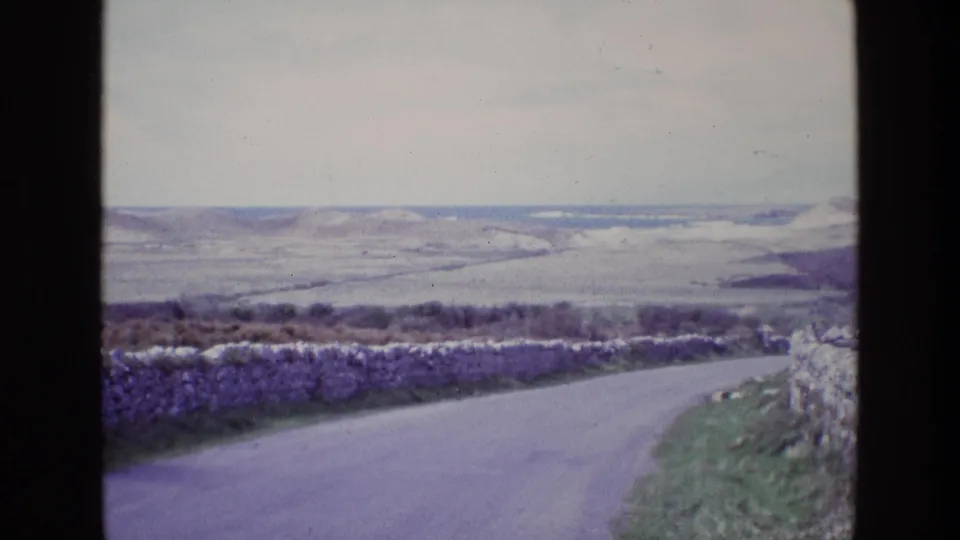 Vintage 8mm view in Ireland showing a quiet rural road, stone boundary wall, grassy verge, and broad hills in the distance.