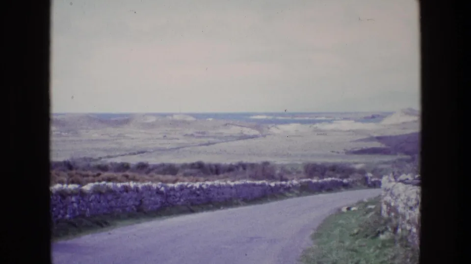 1969 Ireland 8mm frame of a paved country road beside stone walls, with muted hills and fields stretching across the background.