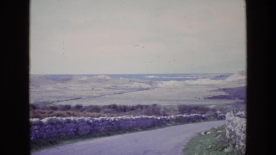 End frame from a 1969 Ireland home movie showing a road edge, stone walls, grassy bank, and distant rolling countryside.
