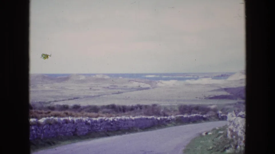 1969 Ireland 8mm view of a coastal road bending past a stone wall toward pale fields, low hills, and a distant water horizon.