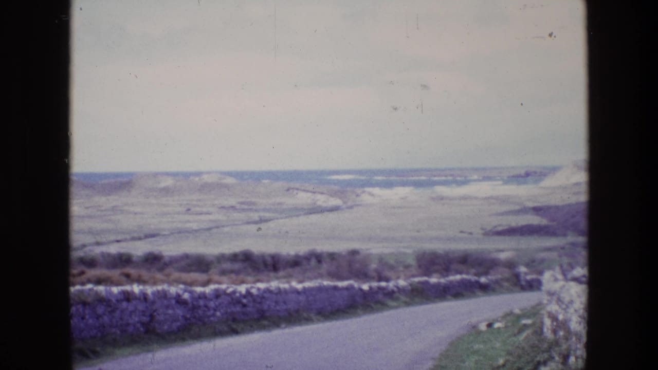 Stone-walled roadside foreground with pale hills and a distant sea horizon beyond.