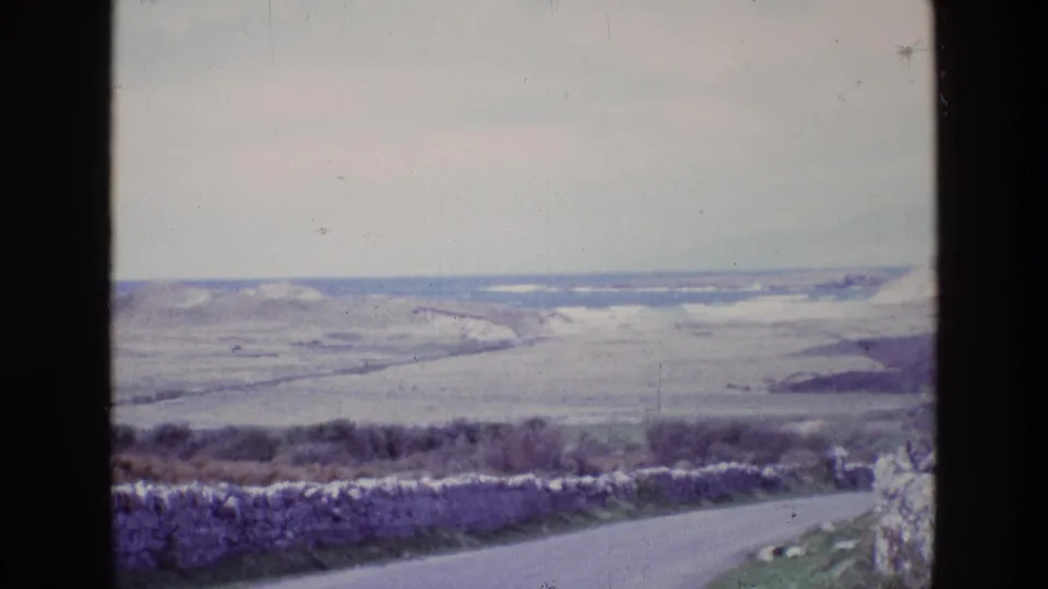 1969 Ireland home-movie frame showing a quiet coastal road, rough stone wall, open fields, low hills, and distant water.