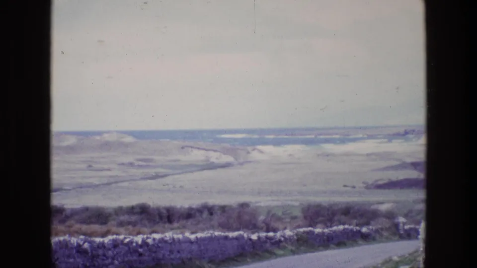 1969 Ireland 8mm coastal outlook over a roadside stone wall, with pale fields, low hills, and a distant waterline.