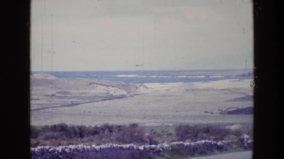 1969 Ireland 8mm travel frame with a hazy shoreline, grassland slopes, and foreground scrub below the window line.