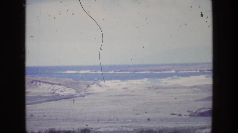 1969 Irish coastline frame with sandy dunes below, blue-gray water beyond, and visible 8mm film wear across the sky.