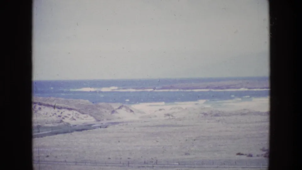 Faded 8mm frame of an Irish coastal panorama, showing pale dunes, open grassland, water, and a distant shoreline.