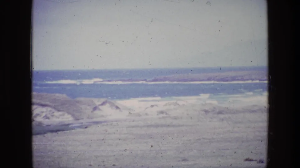 1969 8mm home-movie still of Ireland's coastline, with dunes, muted grassland, surf, and a hazy sea horizon.