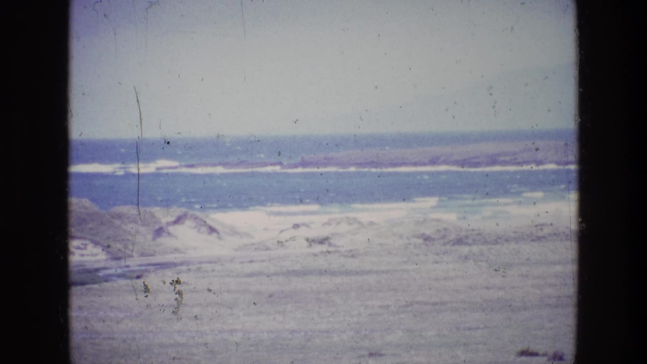 1969 8mm home-movie frame from Ireland showing a pale coastal landscape with water, dunes or beach, and distant low land.