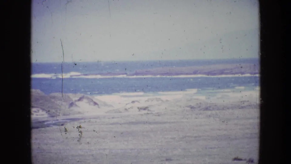 1969 8mm home-movie frame from Ireland showing a pale coastal landscape with water, dunes or beach, and distant low land.