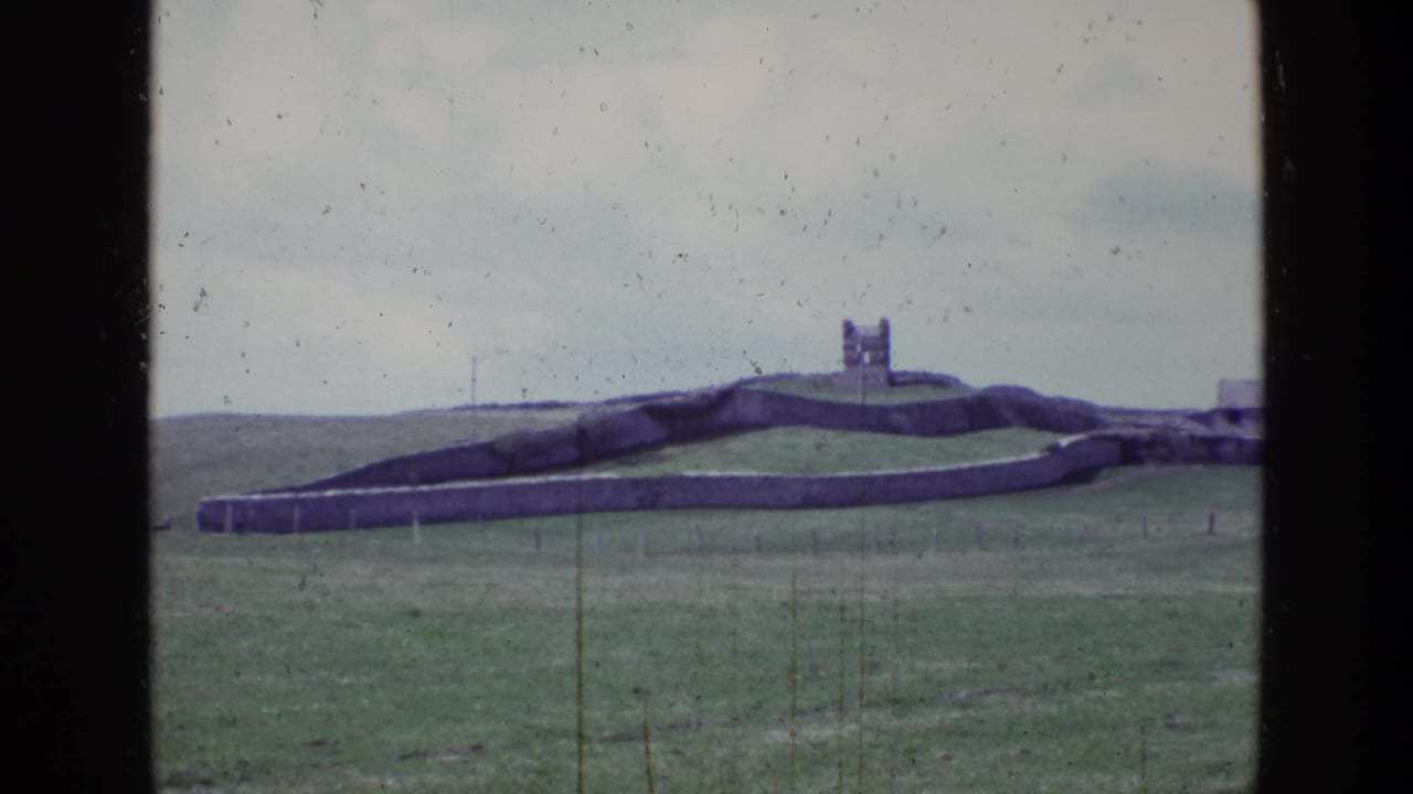 1969 Ireland 8mm frame showing a stone fort or ruin on a green hill, bordered by a long stone wall and open grassland.