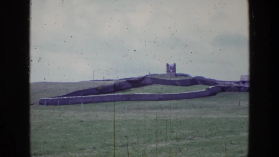 1969 Ireland 8mm frame showing a stone fort or ruin on a green hill, bordered by a long stone wall and open grassland.