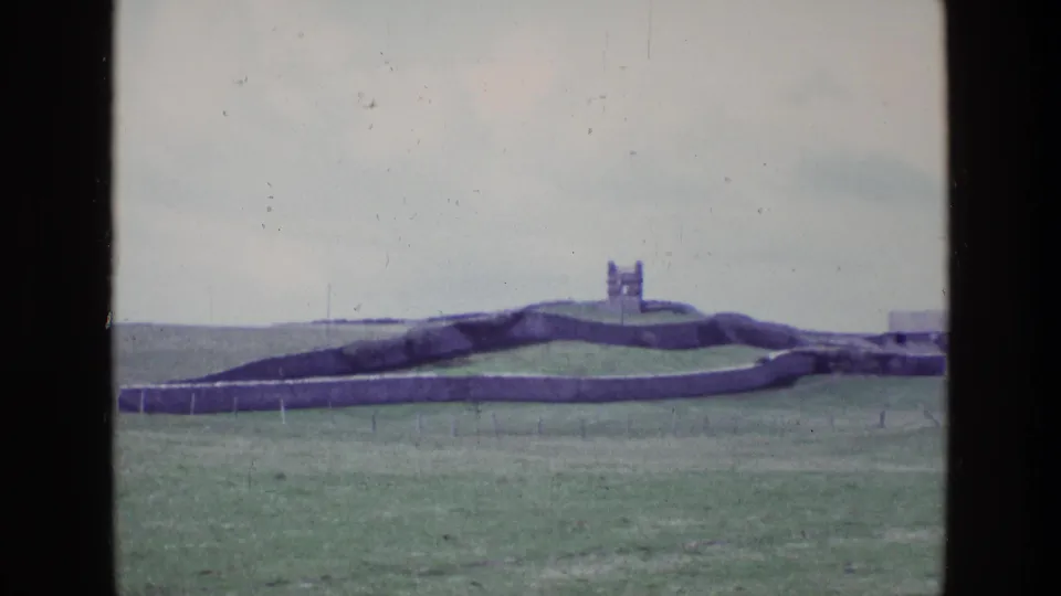 1969 Ireland 8mm frame of a hilltop stone fort and extended wall seen from a steady visitor viewpoint across grassland.