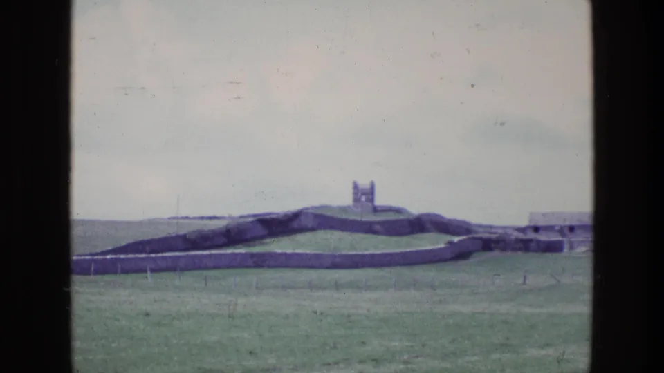 1969 Ireland 8mm frame showing the stone-walled hill fort landscape, with open grass and a low structure visible at the right edge.