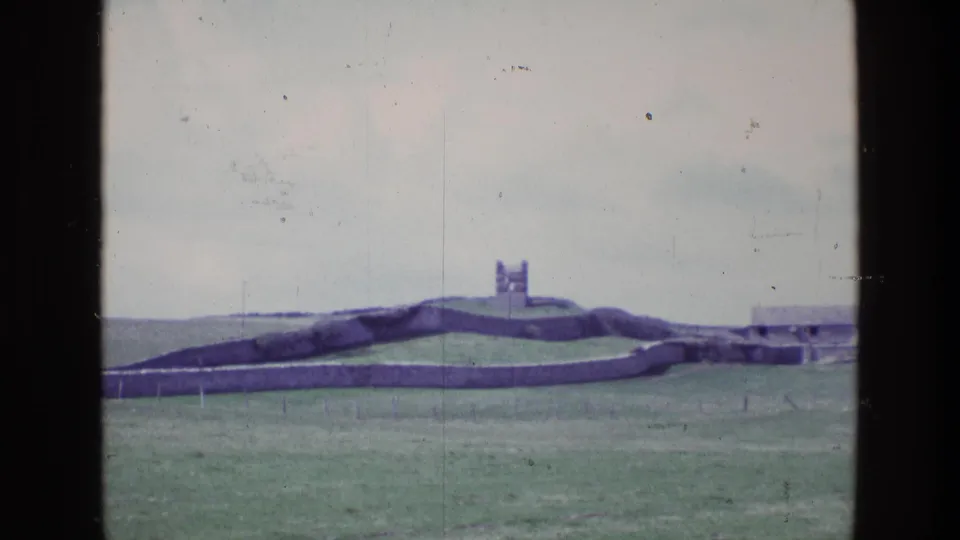 1969 Irish 8mm view of a green field, low stone walls, and a small distant tower under a pale sky.
