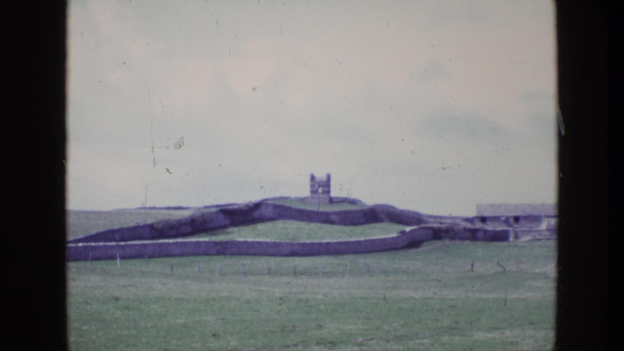 Distant tower beyond rolling green field and stone walls.