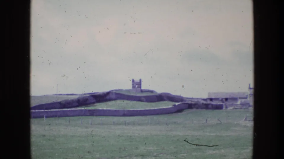 1969 Irish home-movie frame showing a grassy field, long low stone wall, distant tower, and faint fencing.