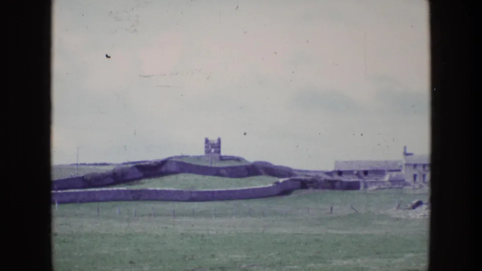 1969 Ireland 8mm frame of green fields, layered stone walls, a distant tower, and low buildings near the right edge.