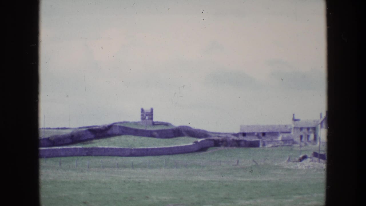 1969 Irish 8mm frame showing a distant fortified tower above grassland, with stone walls, low coastal buildings and pale overcast sky.