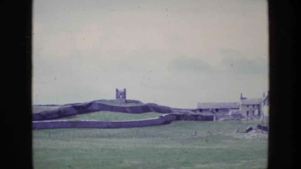 1969 Irish 8mm frame showing a distant fortified tower above grassland, with stone walls, low coastal buildings and pale overcast sky.
