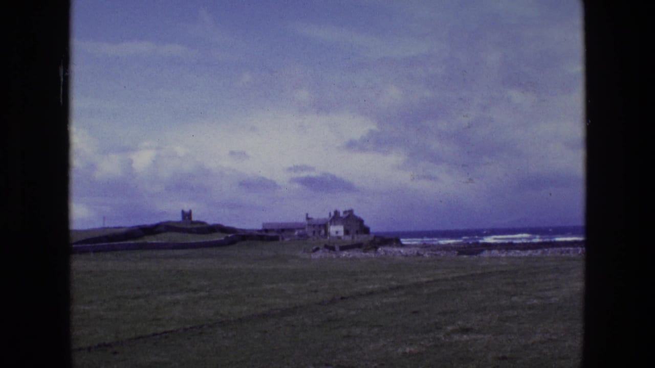 1969 Irish coastal landscape with a tower on a low hill, weathered shoreline buildings, open grass, rocks, sea and heavy clouds.