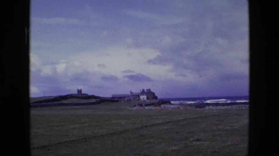 1969 Irish coastal landscape with a tower on a low hill, weathered shoreline buildings, open grass, rocks, sea and heavy clouds.