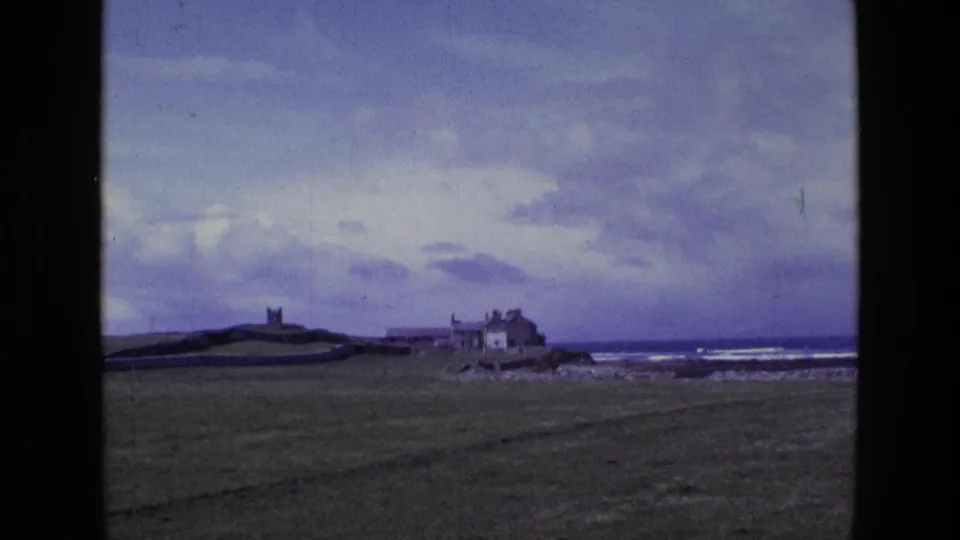 Irish 1969 home-movie frame looking across grassland toward a small tower, stone boundary walls, coastal buildings and a clouded seascape.