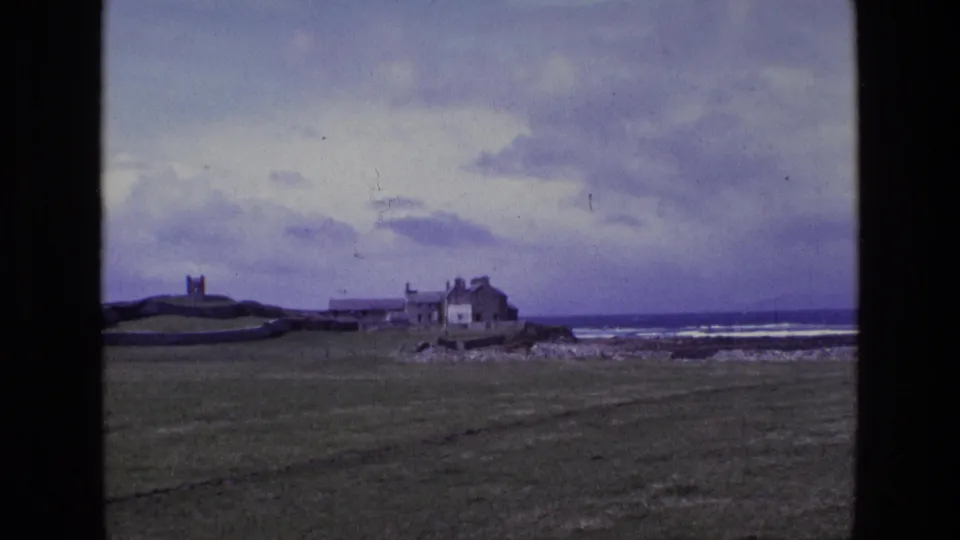 1969 Ireland 8mm frame with a fortified tower on a grassy rise, low shoreline buildings, rocky coast, white surf and overcast sky.