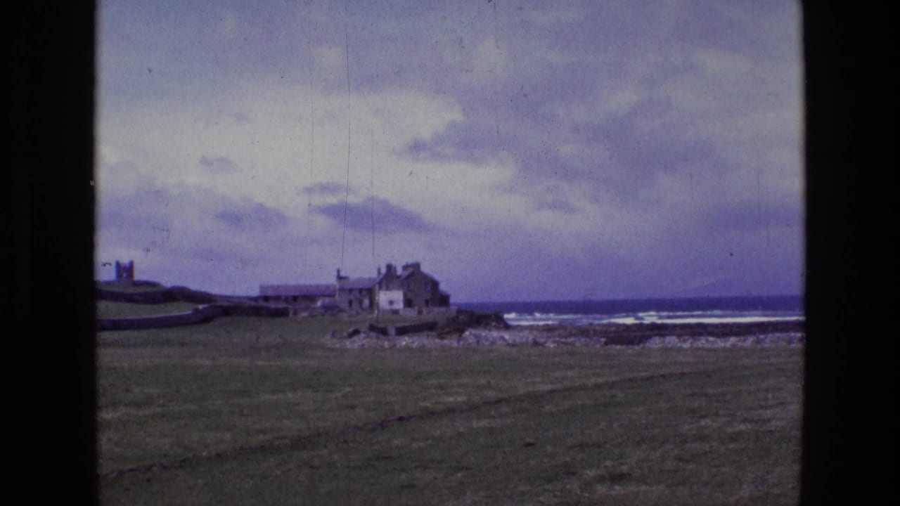1969 Ireland 8mm frame showing a grassy coastal field, rocky shoreline, surf, and a house with outbuildings near the ocean.