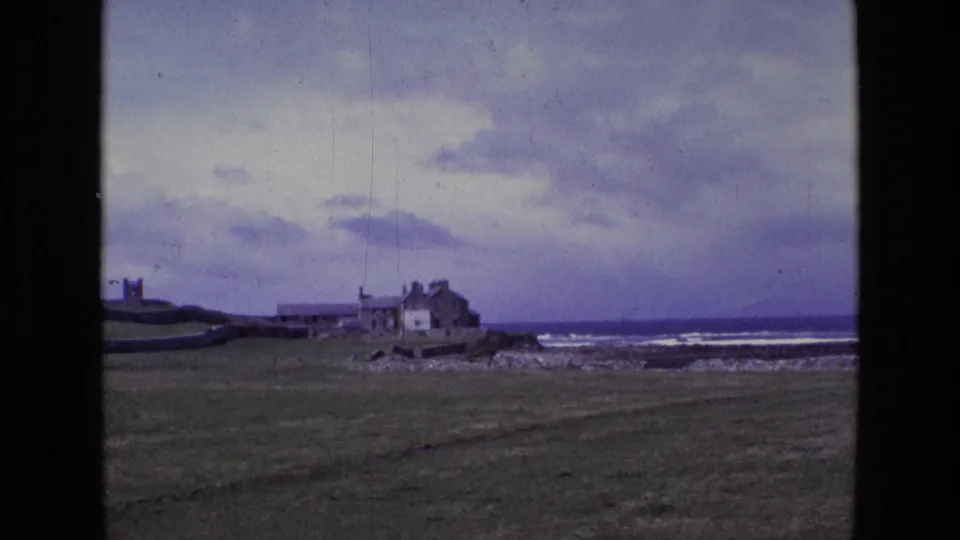 1969 Ireland 8mm frame showing a grassy coastal field, rocky shoreline, surf, and a house with outbuildings near the ocean.