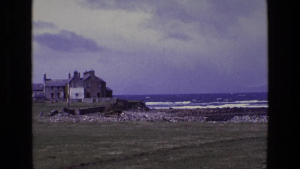 1969 Ireland 8mm frame focused on the shoreline house, grass foreground, rocks, and choppy ocean beyond.