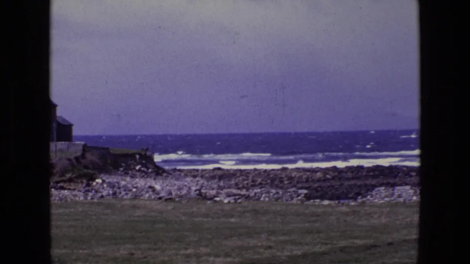 1969 Ireland 8mm frame showing surf, a rocky shore, grass foreground, and the edge of a coastal building after the zoom shifts right.