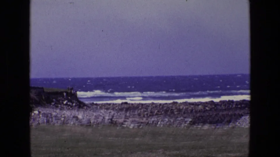 1969 Ireland 8mm frame showing a rocky coast, low grass, rough sea, and white surf with dark film borders.