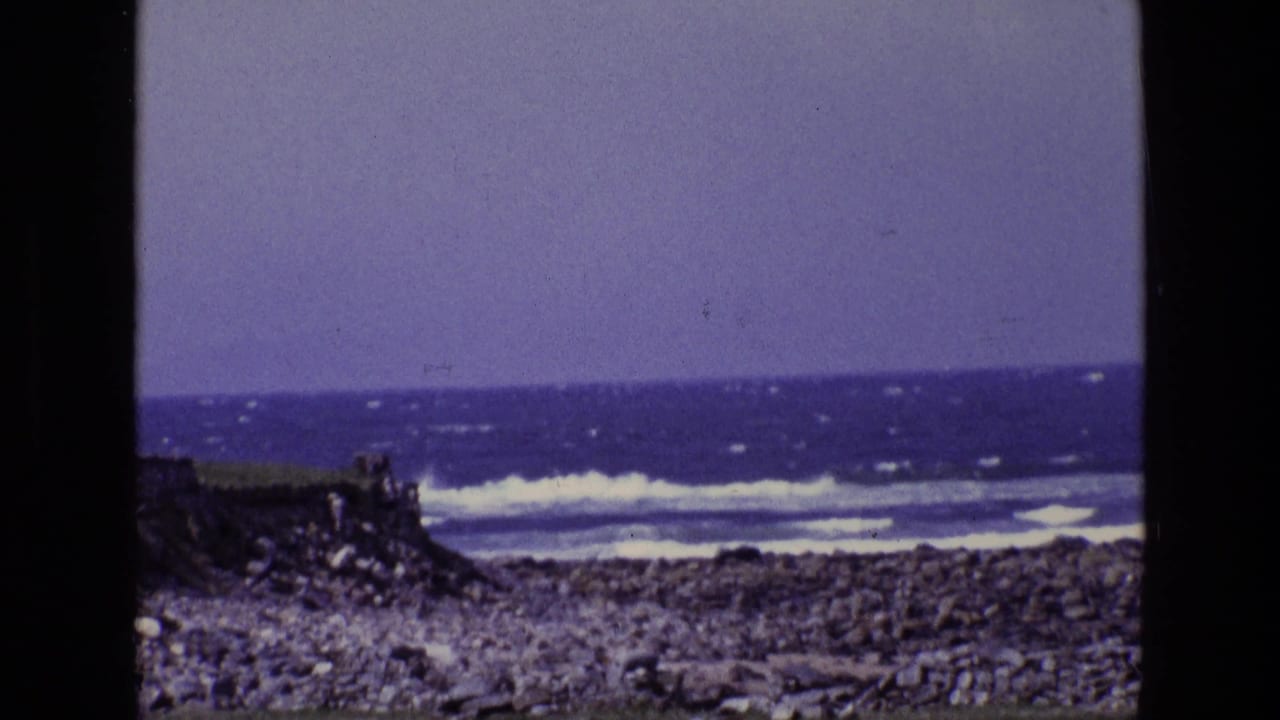 White waves roll behind a rocky coastal foreground beneath a hazy sky.