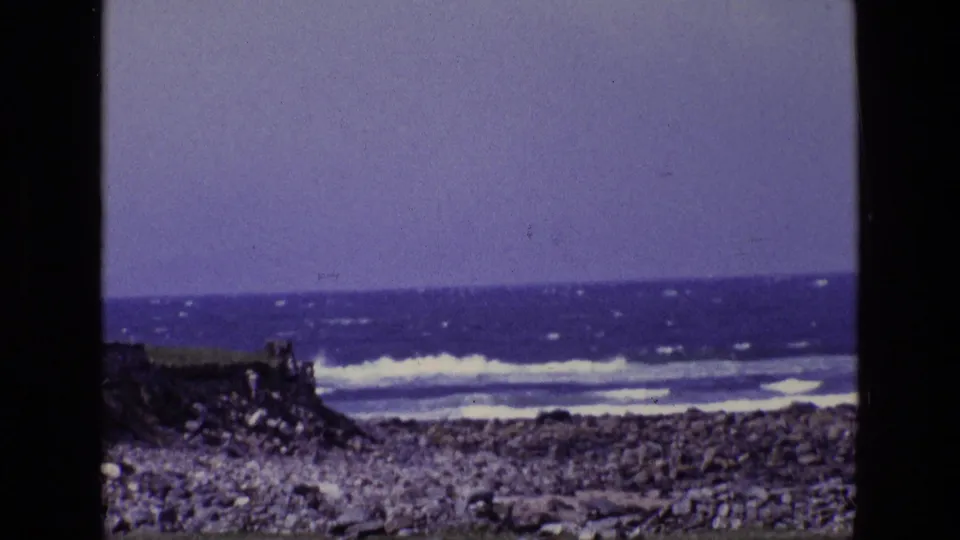 1969 Ireland coastal frame with breaking waves, stone-covered shore, distant horizon, and aged blue-purple film color.