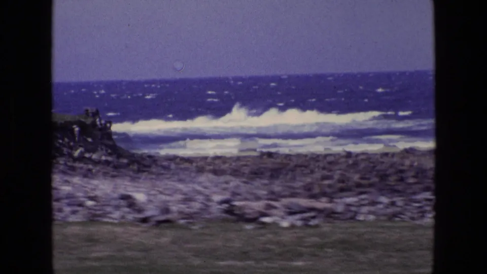 1969 Ireland home-movie frame showing a slightly blurred pan across grass, stones, and choppy coastal surf.