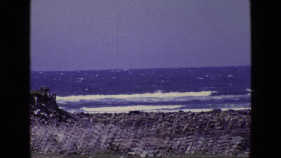1969 Ireland 8mm frame of a stone-covered shoreline, white breaking waves, blue sea, and pale overcast sky.