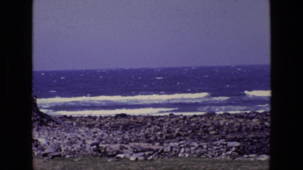 1969 Ireland 8mm frame showing rough ocean waves beyond a rocky coastal shore, with dark film borders and aged purple-blue color.
