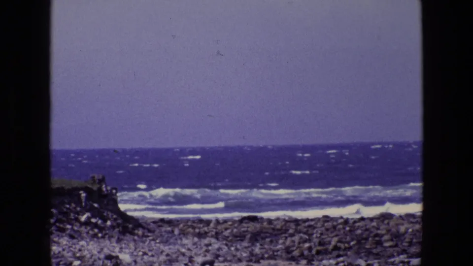 1969 Ireland home-movie frame of surf moving across a rocky shoreline, with dark vegetation at left and visible 8mm grain.