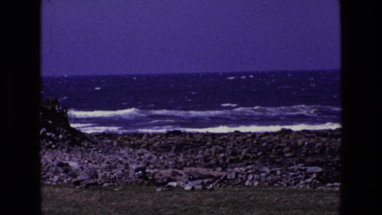 Rocky shoreline with surf, grass foreground, cloudy sky, and dark film borders.