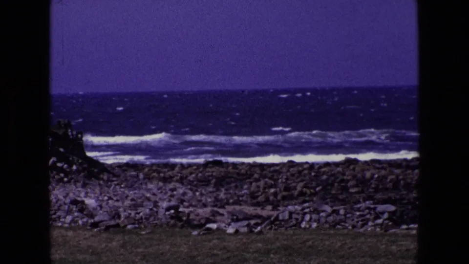 1969 8mm view in Ireland of a rocky coast with breaking waves, a grassy foreground, cloudy sky, and visible film-frame edges.