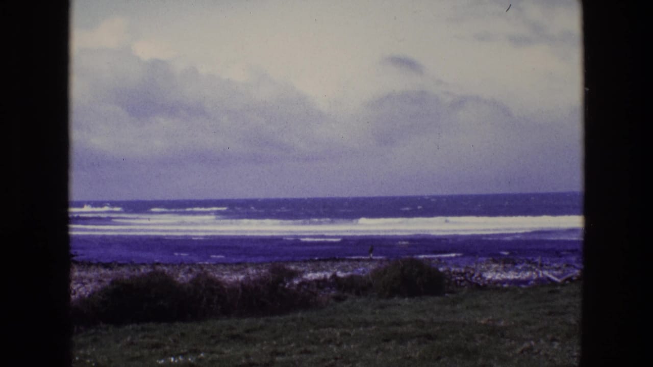 1969 8mm coastal view in Ireland showing cloudy sky, white surf, rocks, grass, and a small distant person near the shoreline.