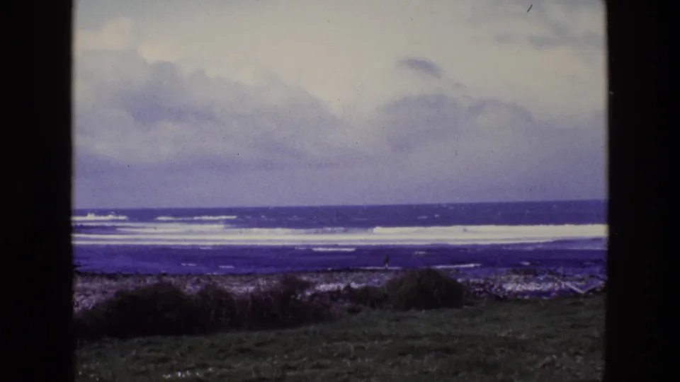 1969 8mm coastal view in Ireland showing cloudy sky, white surf, rocks, grass, and a small distant person near the shoreline.