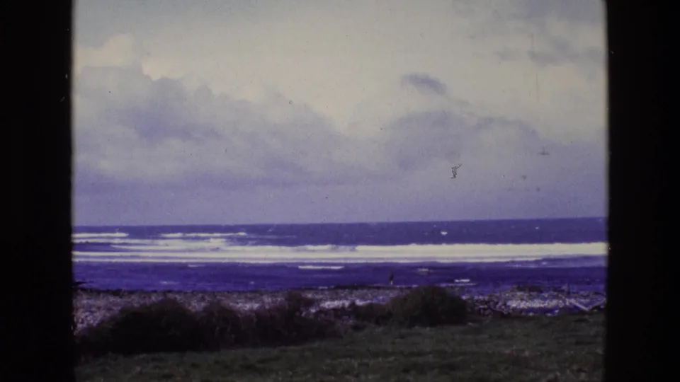 Ireland 1969 home-movie frame with layered surf, overcast clouds, rocky beach, grass in the foreground, and a small distant figure.