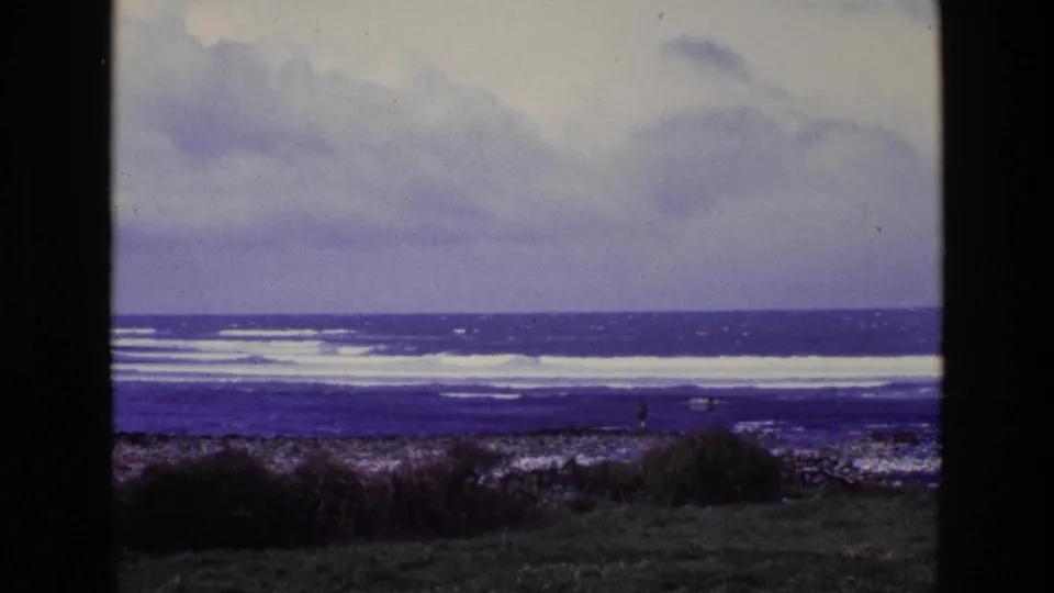 1969 8mm Ireland coastal frame showing white waves, cloudy sky, rocky shoreline, grassy foreground, and a tiny person near the sea.