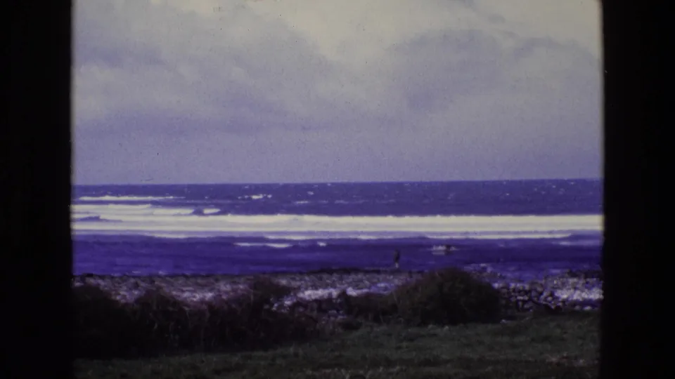 1969 8mm Ireland coastal view with rocky shore, low grass, distant surf, and a small figure near the waterline.