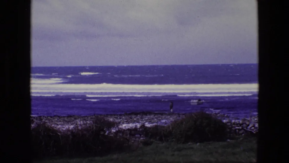 1969 8mm Ireland shoreline scene with strong surf, rocky foreground, distant person, and a small boat near the water.