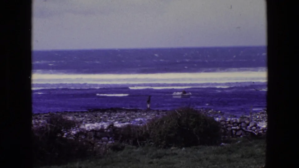 1969 8mm Ireland coast with a distant person on rocks, a small boat in choppy water, and wide bands of ocean surf.